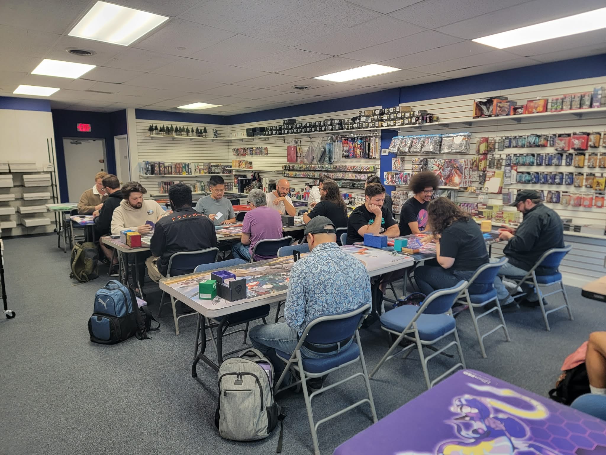 Players at folding tables during a Gundam Card Game tournament, shop shelves of card and game product in the background.