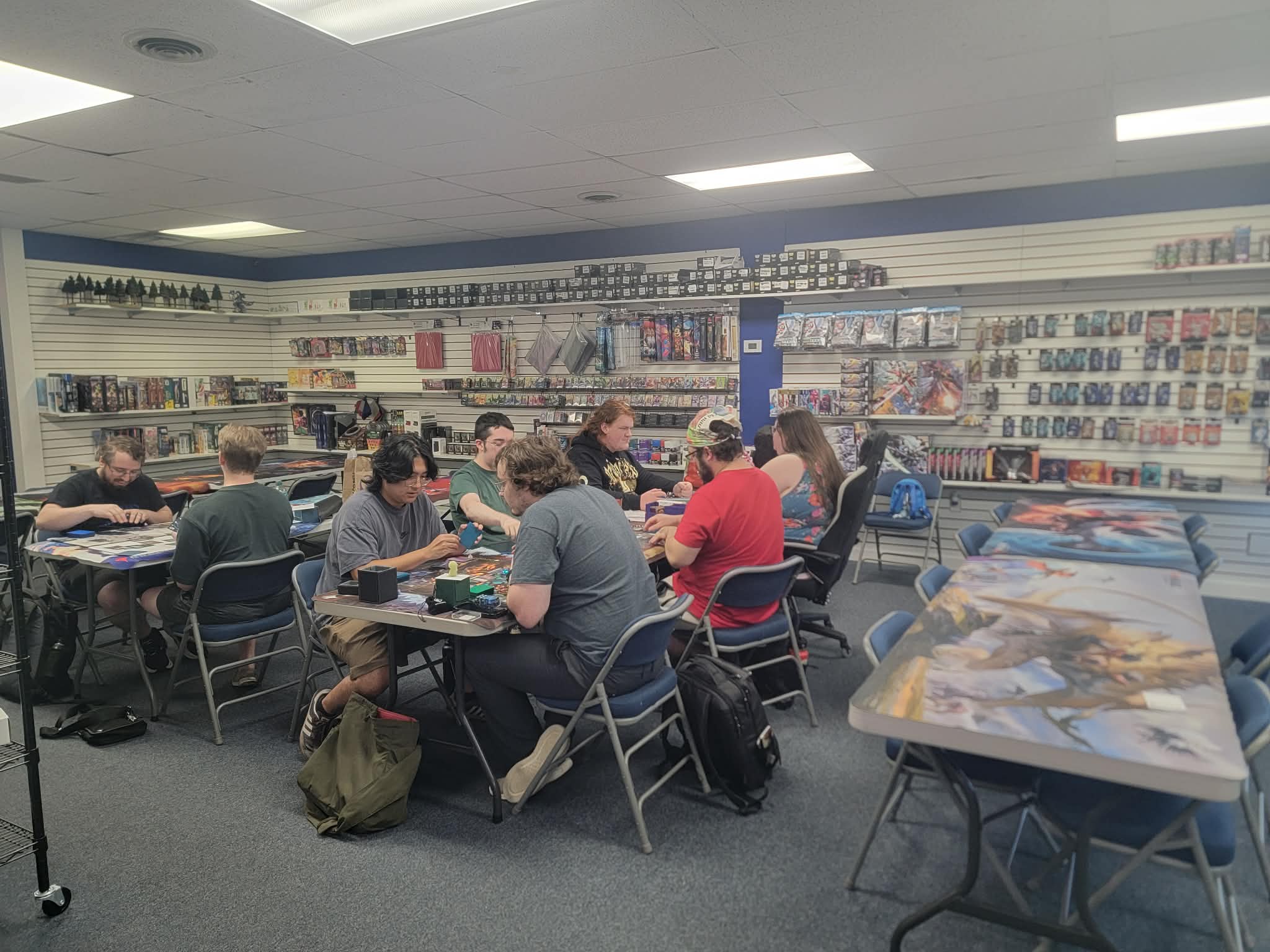 Players seated around folding tables mid-match, shop shelves of card and game product in the background.