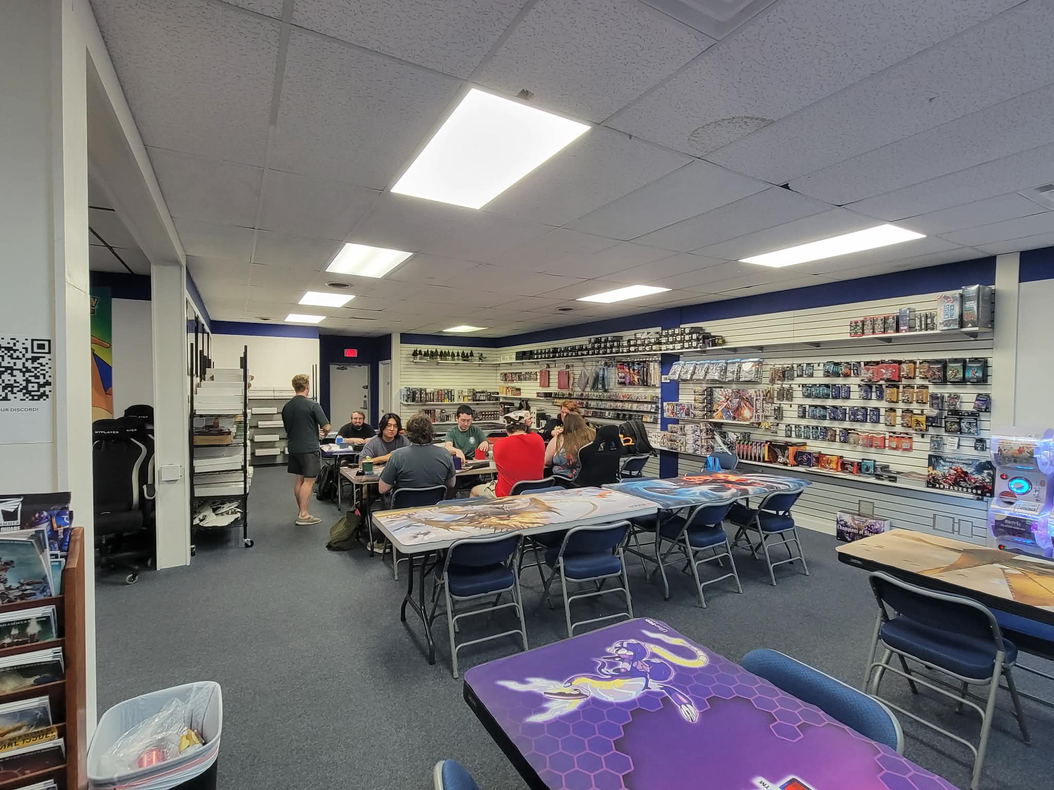 The playgroup at the tables, decks and dice boxes out, with the same wall of product behind them.